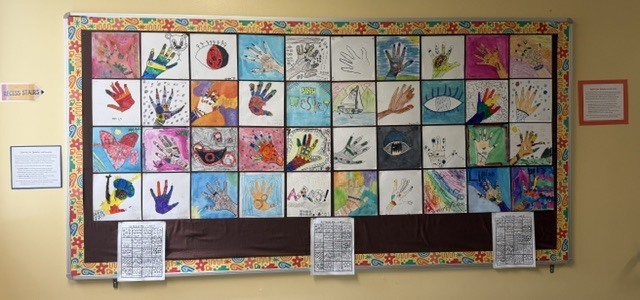 A colorful bulletin board displays numerous handprints in various colors, shapes, and styles. White papers hang below.