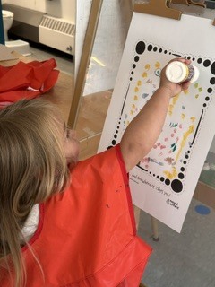 A child wearing a red smock stands at an easel, painting on a large white paper.