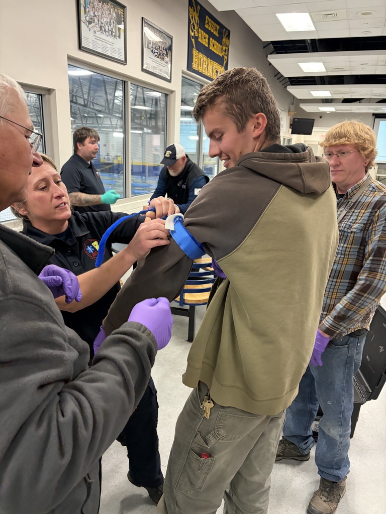 Two individuals in protective gloves assist a man with a device attached to his arm. Four others watch in a hallway with glass walls.