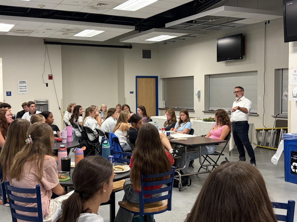 A classroom with students seated at tables, one person standing at a table, and a speaker in the background.