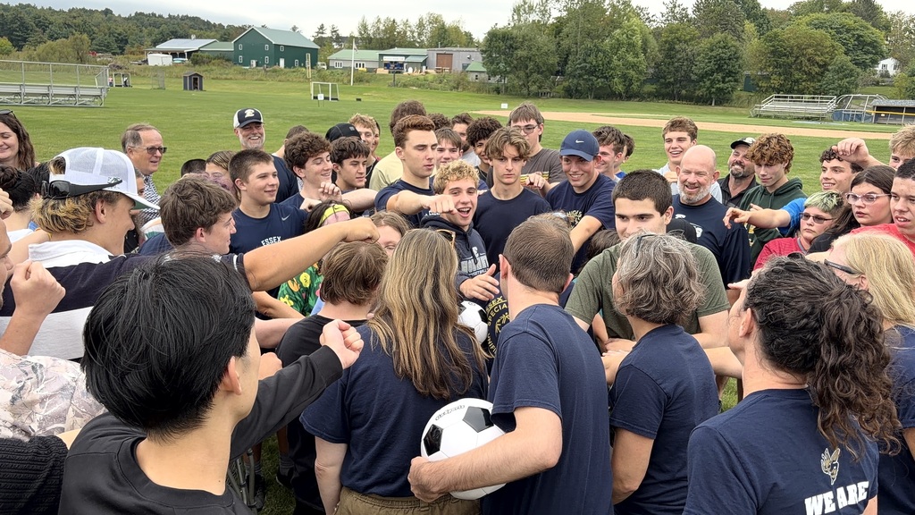 Group of people gathered, some wearing blue shirts, on a field with trees, likely in a huddle.