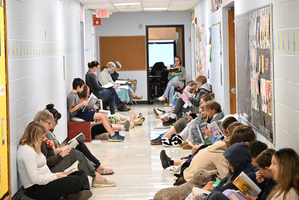 People sit along a hallway, reading books. A door and bulletin boards are on the right.