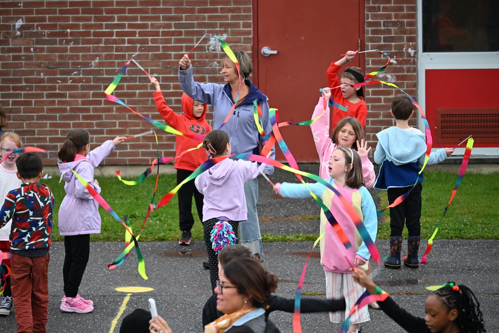 A group of children waving colorful ribbons in an outdoor setting, with a brick building in the background.
