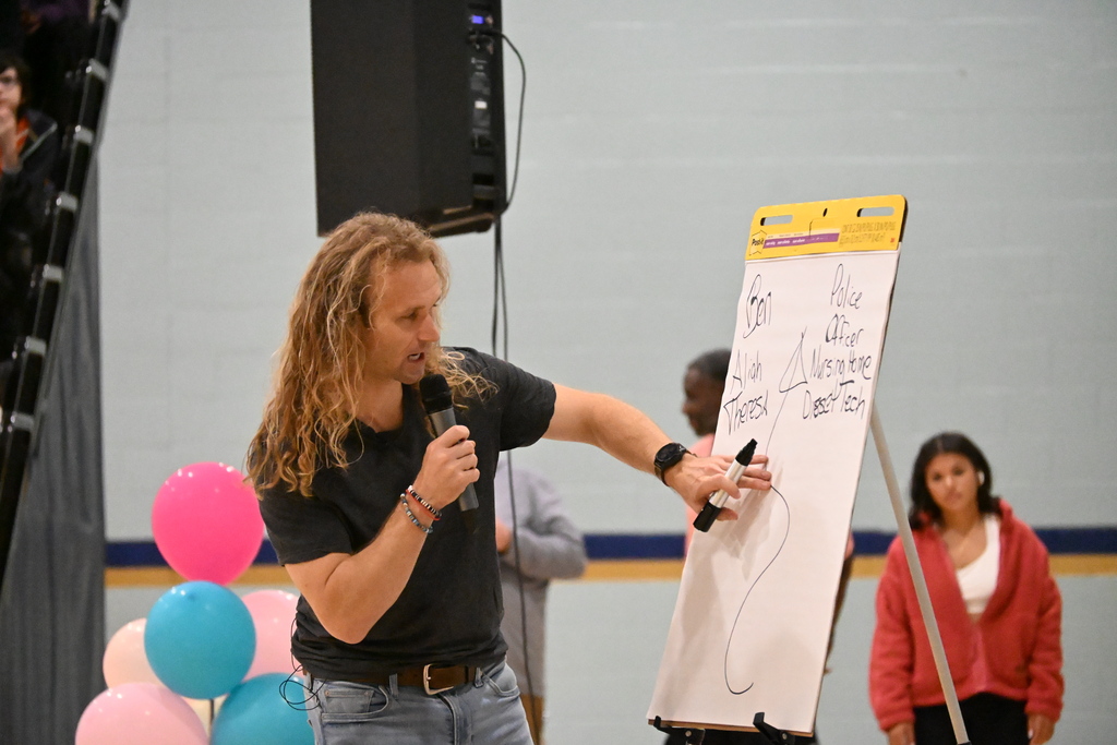 A man with long hair speaks into a microphone while drawing on a whiteboard. A woman in a red jacket stands behind him.