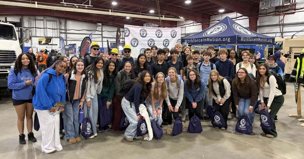 Group of people in a warehouse, wearing various jackets and holding bags, posing for a photo.