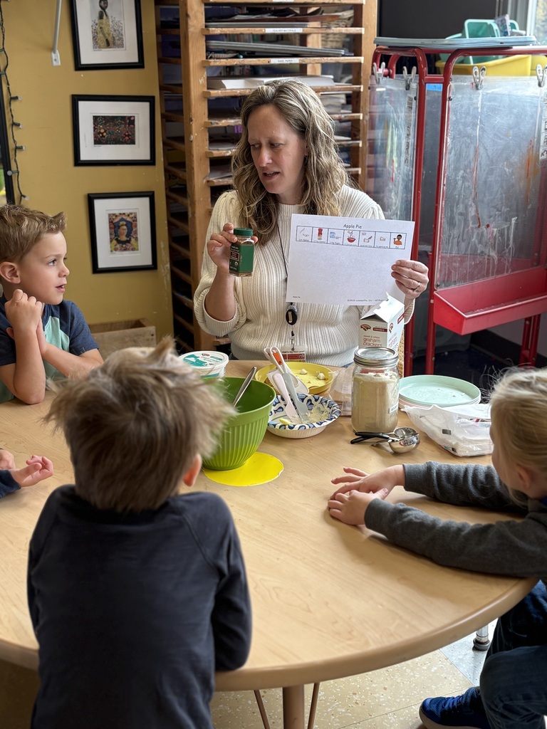 A woman holds a paper while teaching four children at a table. Behind her are shelves, framed pictures, and a red storage unit.
