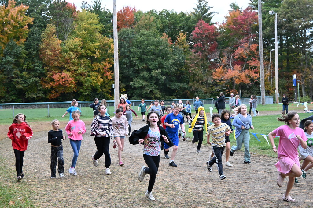 A group of children in various shirts run on a field with dirt ground, colorful trees in the background.