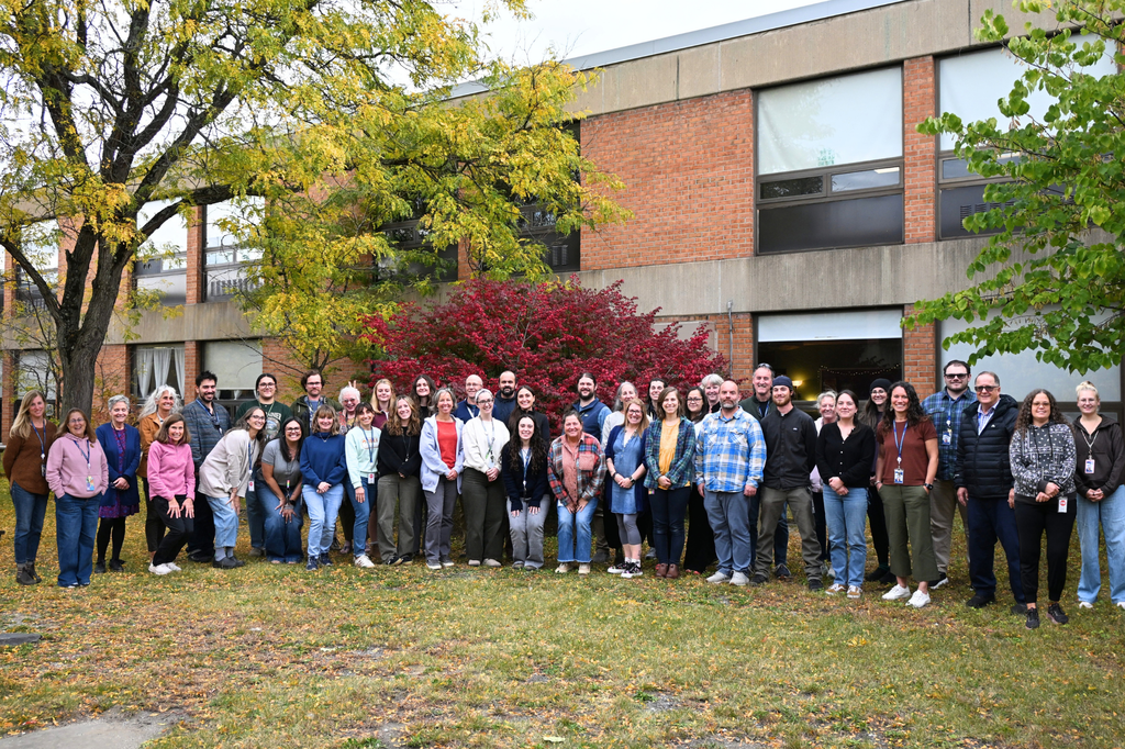 A group of people poses in front of a brick building with trees and plants in the background.