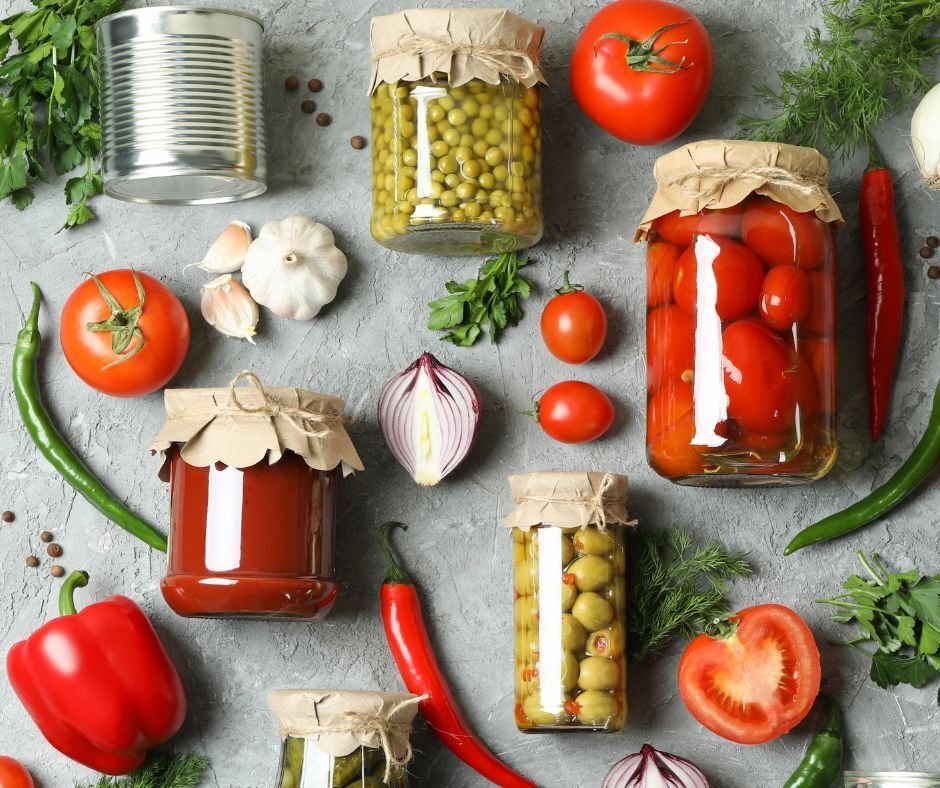 Canned and preserved fruits and vegetables on a gray surface with herbs and spices.