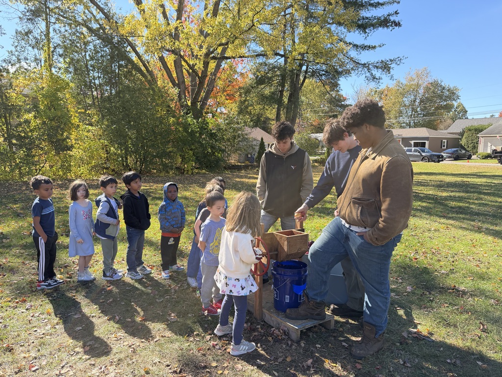 People gathered around a man working on a machine. Trees and houses are visible in the background.