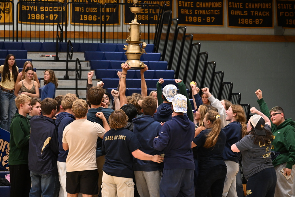 Students and adults in blue and white jerseys raise a trophy as others cheer in the background.