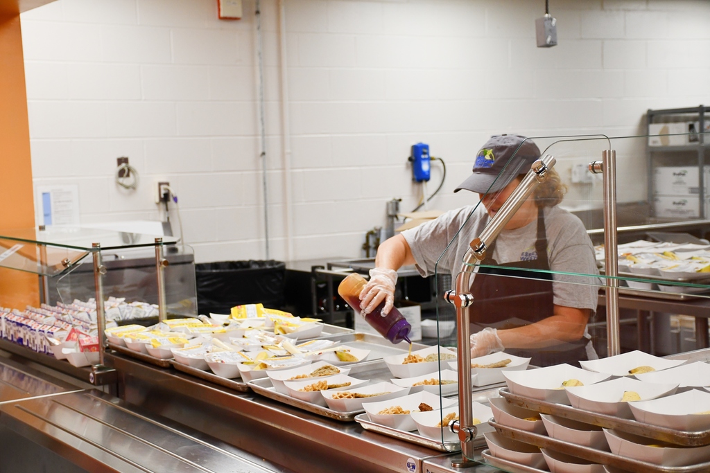 Person wearing hat and apron behind a glass counter, preparing food in trays, with containers and bins in background.