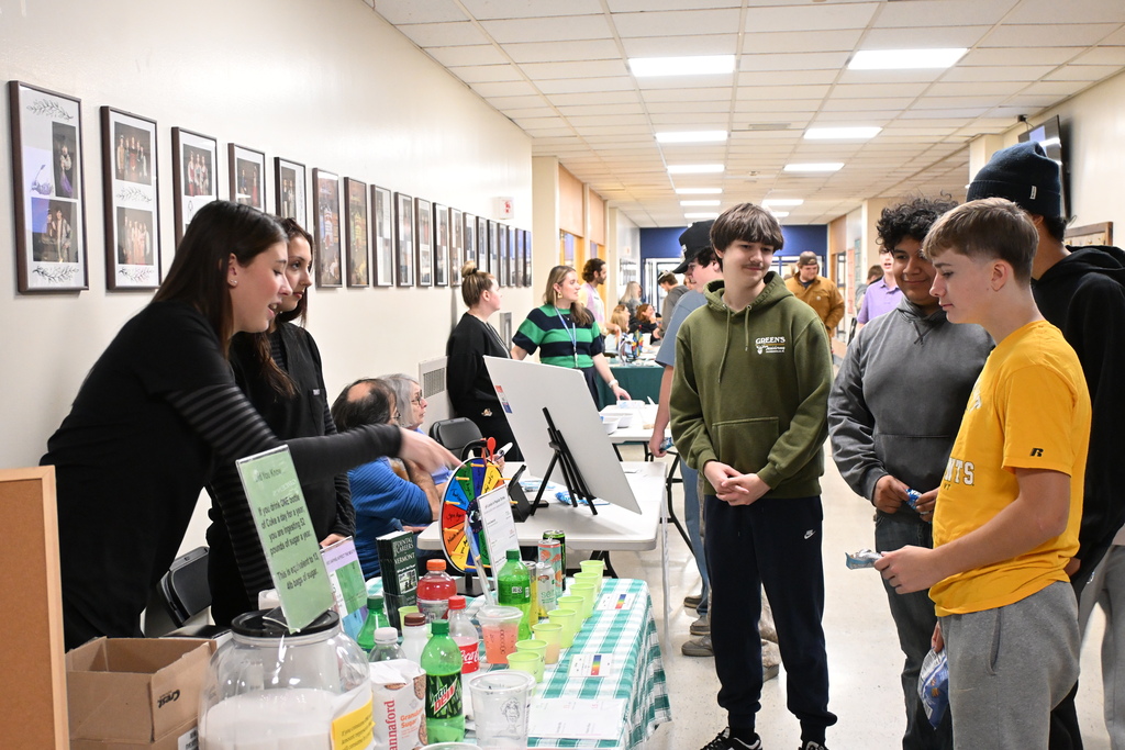 Several students and staff gather around a table with bottles and cups on it in a hallway.
