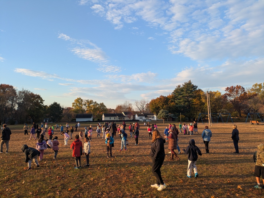 Large group of people standing in an open field with trees, some with jackets, and cloudy sky.