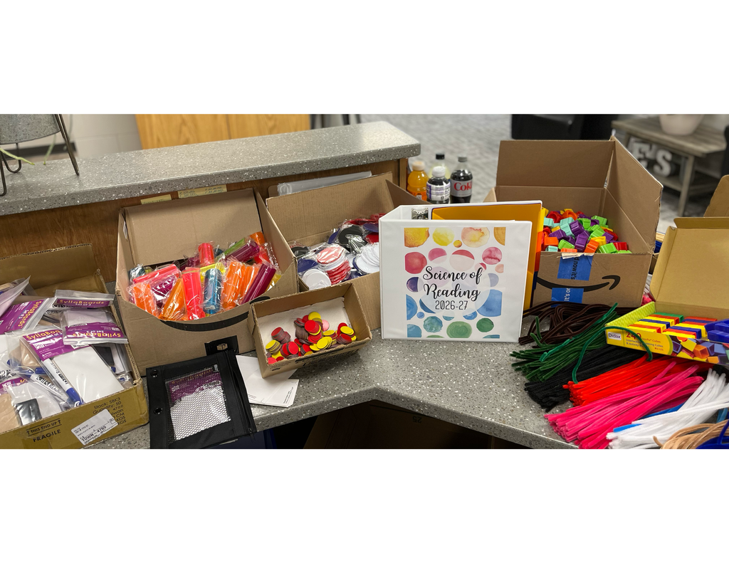 Subject: A high-angle shot of a gray, textured countertop cluttered with various educational manipulatives and workshop materials organized in cardboard shipping boxes and laid out on the surface.  Central Focus:  A white three-ring binder stands upright in the center. The front cover features a colorful design of watercolor-style circles in yellow, pink, blue, and green, with the cursive text: "Science of Reading 2026-27."  Left Side of the Counter:  An open cardboard box containing small, transparent plastic bags filled with brightly colored plastic tubes (orange, pink, and red).  A smaller cardboard tray filled with round, double-sided yellow and red tokens.  Another open box behind it holding stacks of round white and blue plastic tokens.  To the far left, a low cardboard tray holds packaged items labeled "Syllaboards."  A black mesh zipper pouch lies flat on the counter.  Right Side of the Counter:  A large cardboard box filled with small green, purple, and orange plastic items.  A yellow box containing "Linking Cubes" (multicolored connecting blocks).  Large, organized piles of colorful chenille stems (pipe cleaners) in green, brown, red, pink, and white laid out in neat fans directly on the counter.