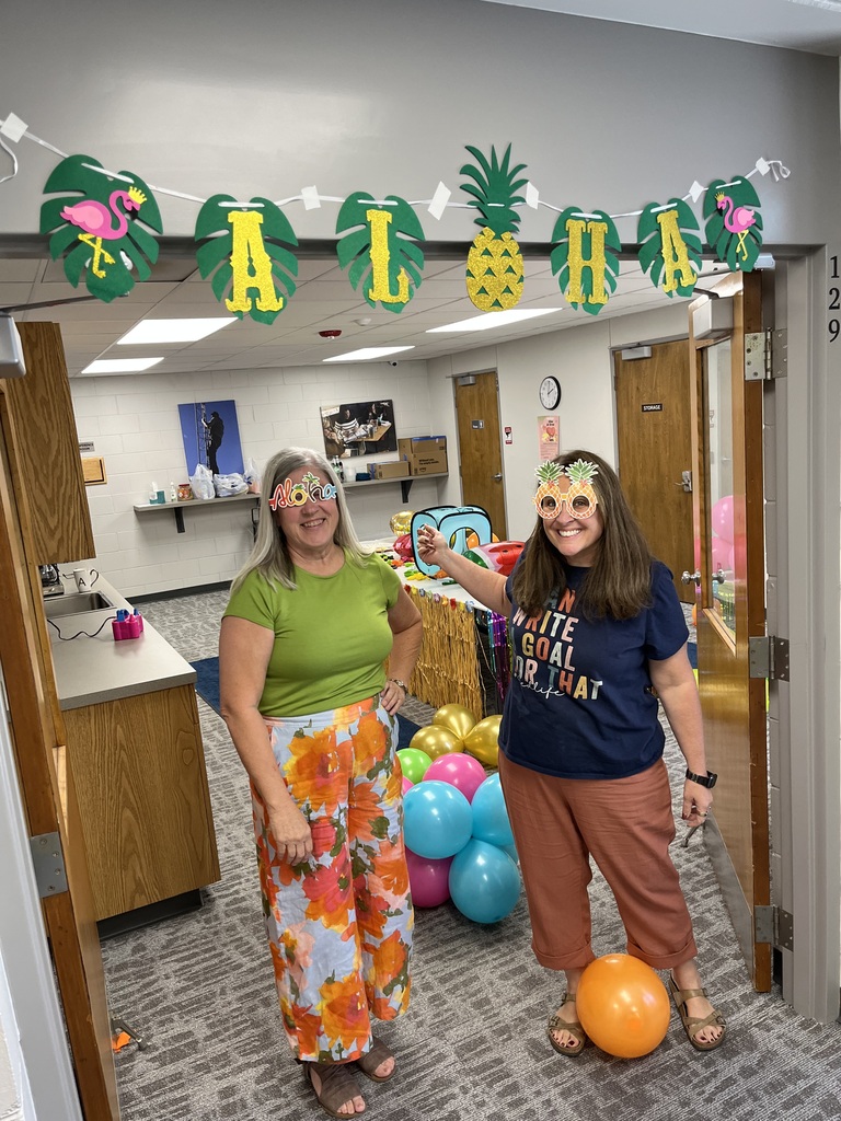 Two smiling women stand in a doorway under a green palm leaf "ALOHA" banner, wearing festive tropical glasses and colorful outfits surrounded by bright balloons.