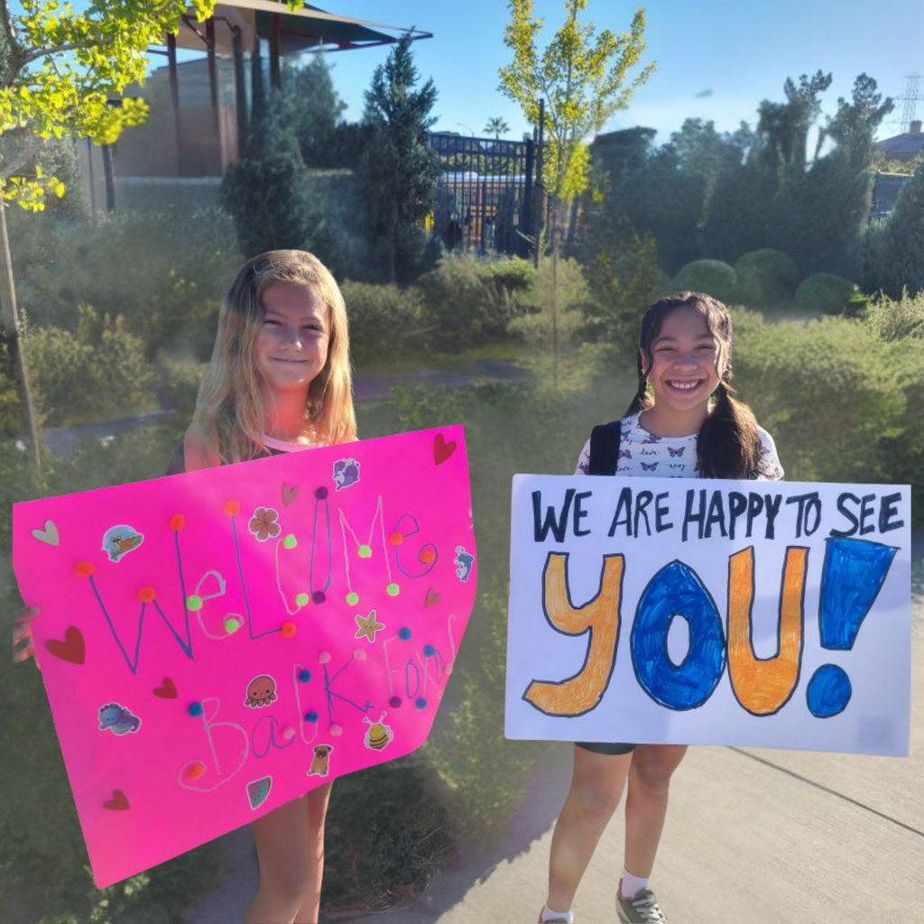Text: We are happy to see you! Welcome Back Image: two female students holding signs.