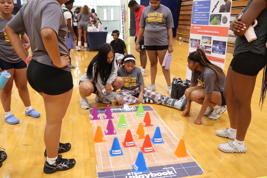 Volleyball players and coach use a robot drone to program through a basketball court maze. A second photo shows Corey Russell and Dr. Ariel Hayes talking with a community member.