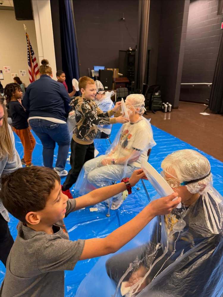 Arbor students pushing plates filled with whipped cream into the faces of teachers who are wearing goggles, ponchos and hair coverings.