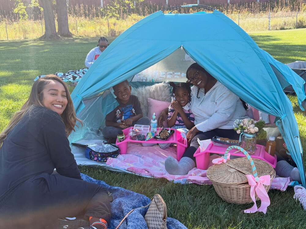 Two children and an adult smile inside a blue tent during an outdoor picnic, while another adult sits beside them on a blanket. Pink trays hold food and drinks, with a wicker picnic basket nearby.