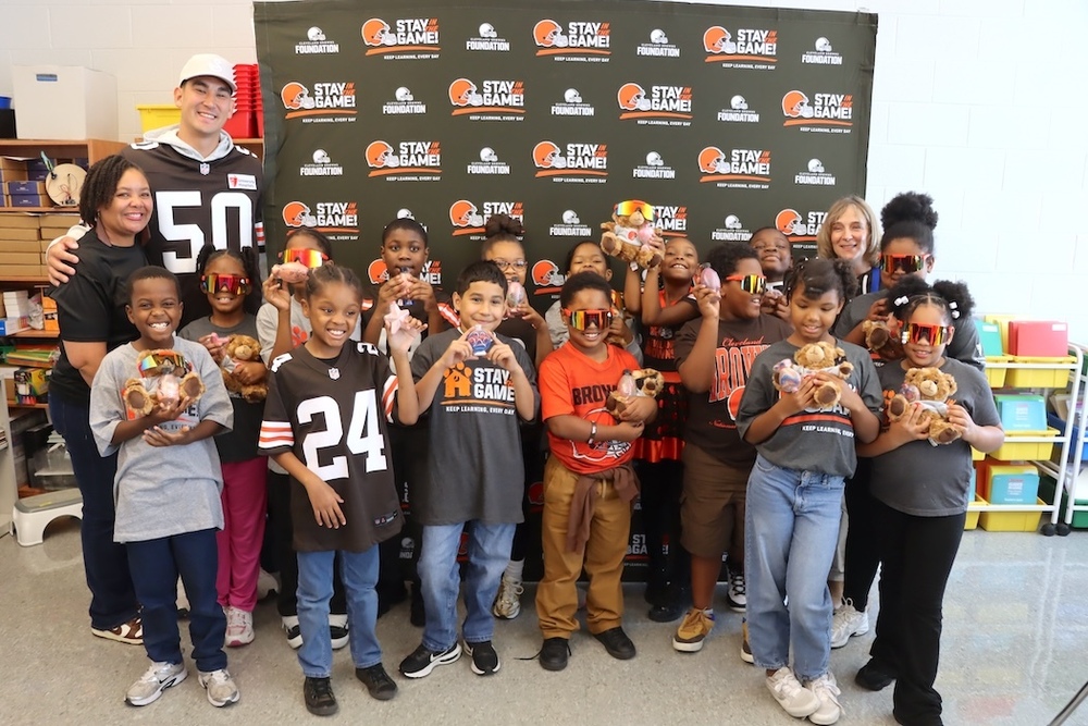 Students and staff pose with a Cleveland Browns player Rex Sunahara in front of a Stay in the Game! backdrop. Students display stuffed dogs and small gifts, and several wear colorful sunglasses and Browns apparel.
