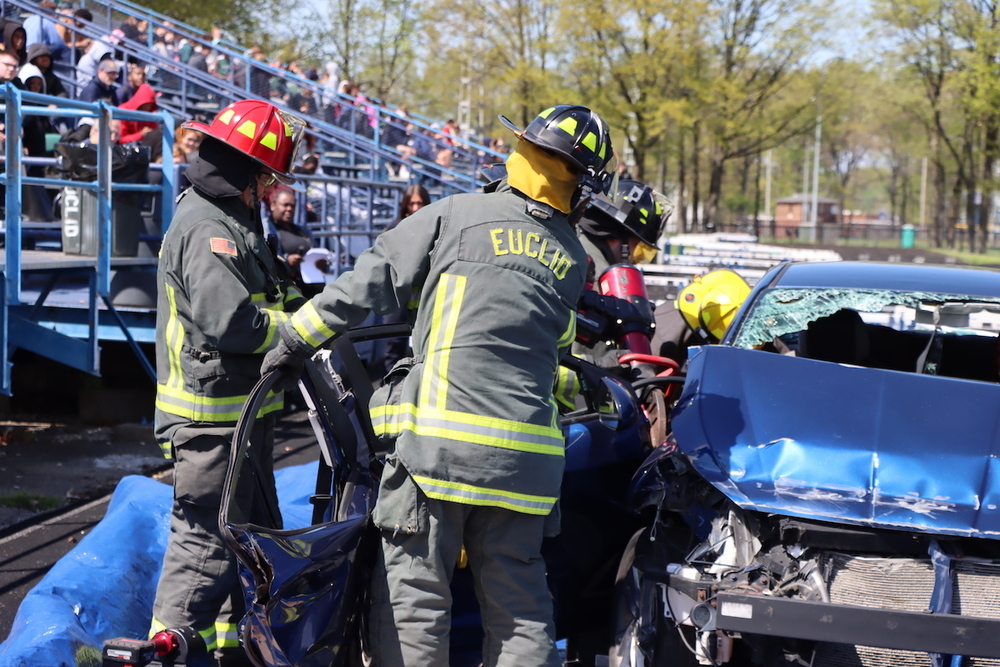 Euclid firefighters use rescue tools to remove a car door during a simulated crash demonstration, with students watching from the bleachers as part of a safety awareness event.