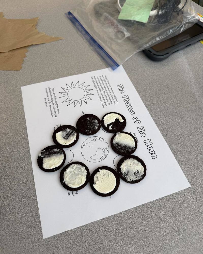 A classroom activity shows Oreo cookies arranged in a circle on a "Phases of the Moon" worksheet, with the cream filling representing the different lunar phases as part of a hands-on science lesson.
