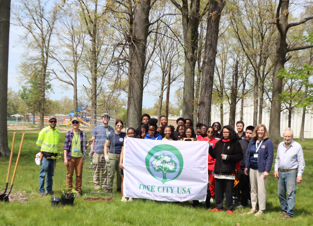 Students and staff gather in a park holding a “Tree City USA” banner during a tree planting event, surrounded by trees, shovels, and equipment on a sunny day.