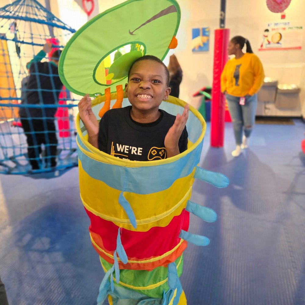 A student enthusiastically plays inside a colorful tunnel structure at We Rock the Spectrum, smiling and engaging in a fun, interactive learning or recreation space alongside classmates and staff.