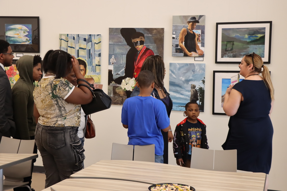 Patrons of the Art Show looking at some of the student artwork that was hung in the Culinary Bistro. There are six different paintings depicting fish, self portraits and a landscape of a river.