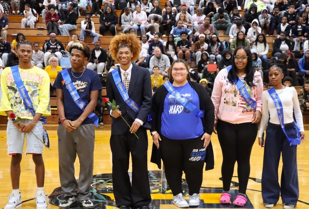 Six Euclid High School Homecoming King & Queen candidates wearing blue sashes pose on the gym floor, with students filling the bleachers behind them. Two court members hold red roses.