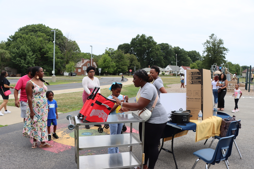 People stand near a table with boxes, chairs, and a cart. One woman hands out a bag. Trees and houses are in the background.