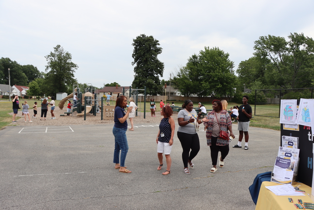 A group of people in an outdoor area, possibly a park, with a playground, tables with materials, and a clear sky.