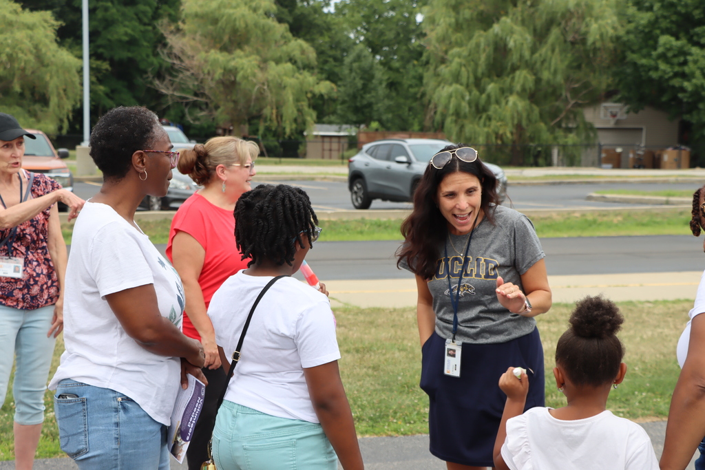 Five women and a child converse near a parking lot. A woman wears a gray shirt. Vehicles drive in the background.