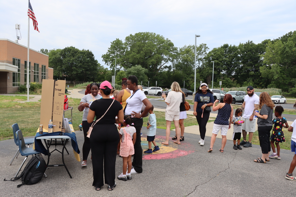 People gather outside near a flagpole. They stand in groups around a table and a chalkboard on the ground.