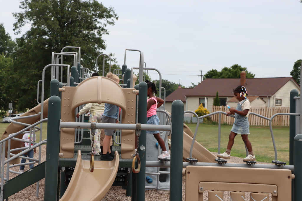 Children at a playground with slides and climbing structures, surrounded by trees and a house.