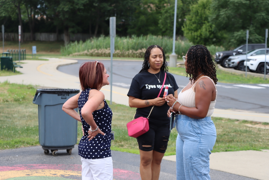 Three women converse on a pathway. A trash bin and greenery are on the left. Trees and parked cars are in the background.