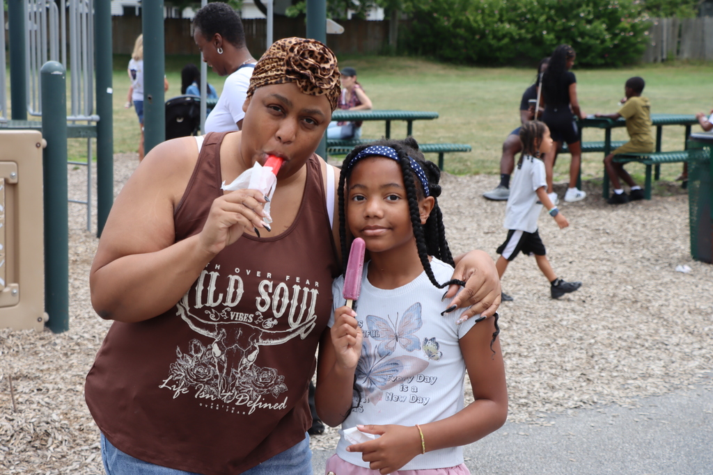 A woman and a child pose with popsicles at a playground. Other people are in the background.