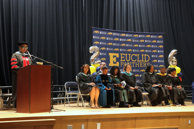 Dr. Ariel Hayes speaking at a podium, while Euclid Schools Administrators and Board Members sit on stage in front of a Euclid Schools step and repeat.