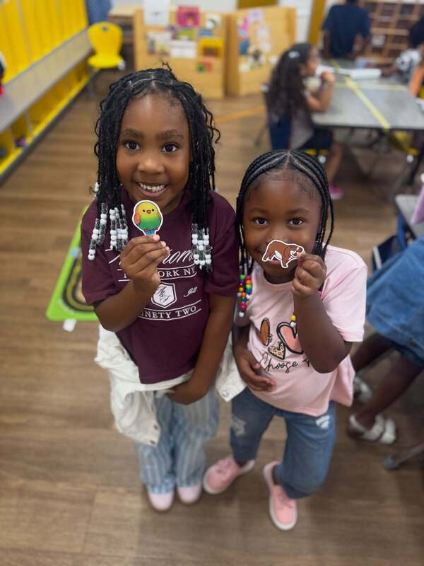 Two students smile at the camera in an Early Learning Village classroom, one holding a colorful bird sticker and the other holding a bulldog sticker.