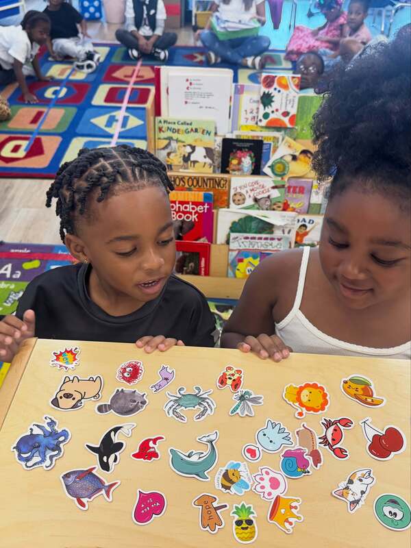 Two Early Learning Village students look over colorful animal and character stickers spread across a classroom table, with books and classmates in the background.