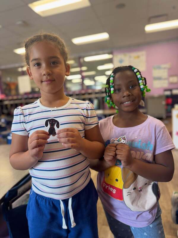 Two Early Learning Village students pose together in a classroom, one holding a dachshund sticker and the other holding a unicorn sticker.