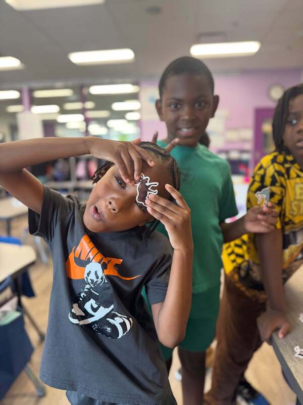 An Early Learning Village student holds an animal sticker against their forehead while two classmates pose behind them, one smiling and making a peace sign.