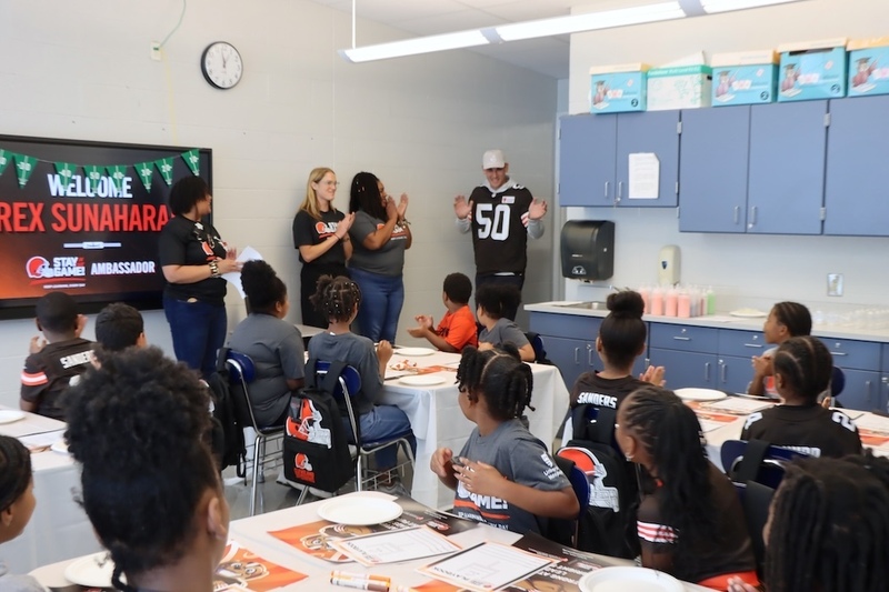 Cleveland Browns player Rex Sunahara stands at the front of a Bluestone Elementary classroom as students and staff applaud. A screen reads, “Welcome Rex Sunahara” and identifies him as a Stay in the Game! ambassador.