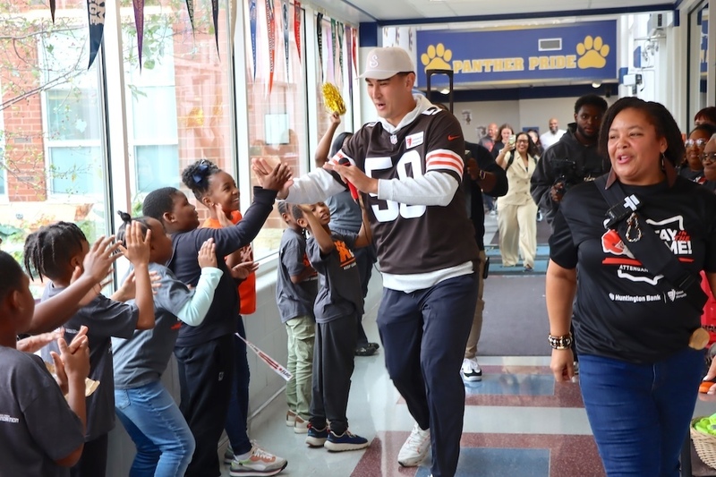 Cleveland Browns player Rex Sunahara high-fives students lining a Bluestone Elementary hallway as staff walk alongside him. A navy and gold “Panther Pride” banner hangs in the background.