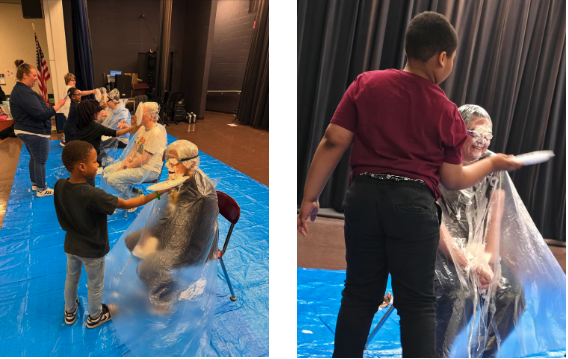 Students participate in a playful school event, throwing whipped cream pies at staff members seated on chairs and covered in ponchos, with smiles and laughter as the activity unfolds on a tarp-covered stage.