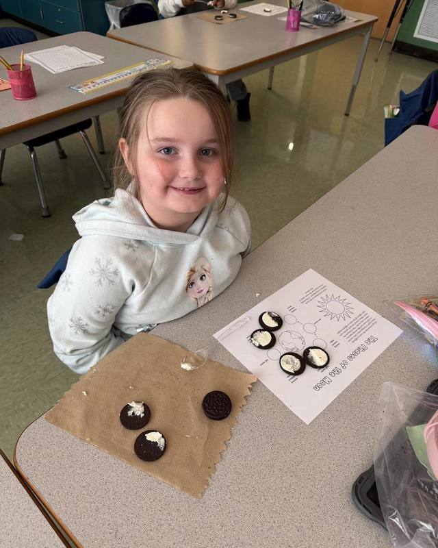 A student sits at her desk smiling beside a worksheet on the phases of the moon, using chocolate sandwich cookies to represent different lunar phases during a classroom science lesson.