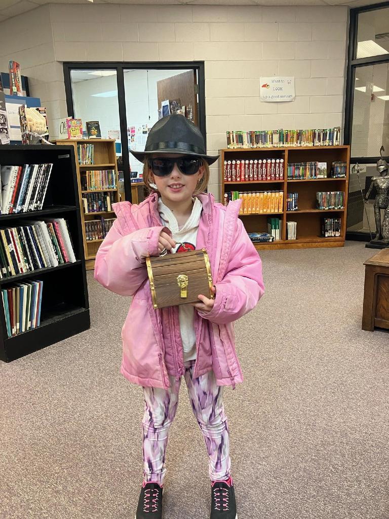 Elementary girl in pink coat, black hat, and black sunglasses holding a small wooden treasure chest