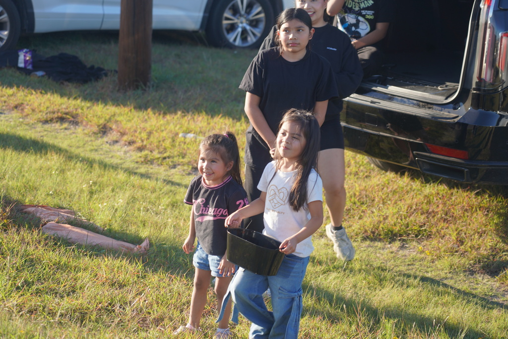 Four children walk on grass. One child holds a black bucket. One child lies on ground.
