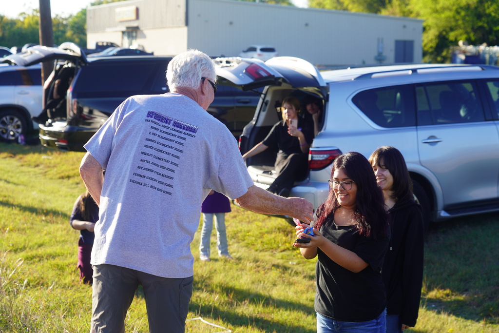 A man in glasses gives a young woman a small object. Other people are standing behind them. Cars are parked in a grassy area.